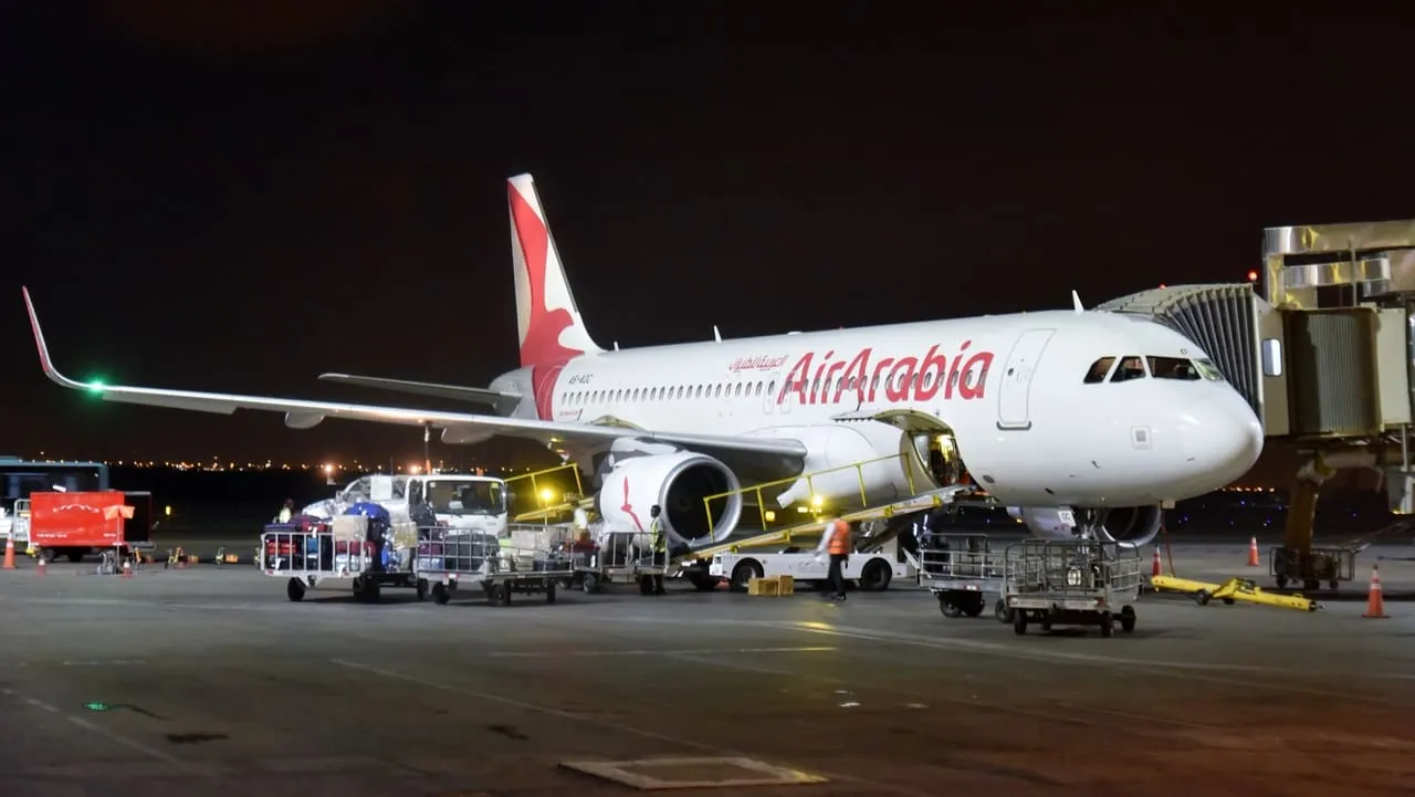 An Air Arabia Airbus A320 parked at an air bridge at Sharjah International Airport at night, with baggage carts beside it