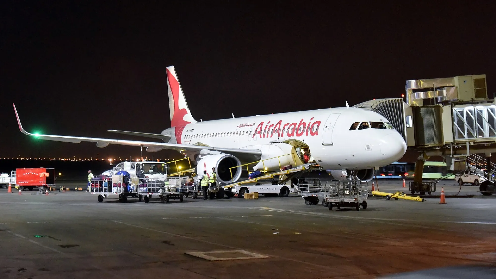 An Air Arabia Airbus A320 at a jet bridge at Sharjah airport at night, with baggage being unloaded