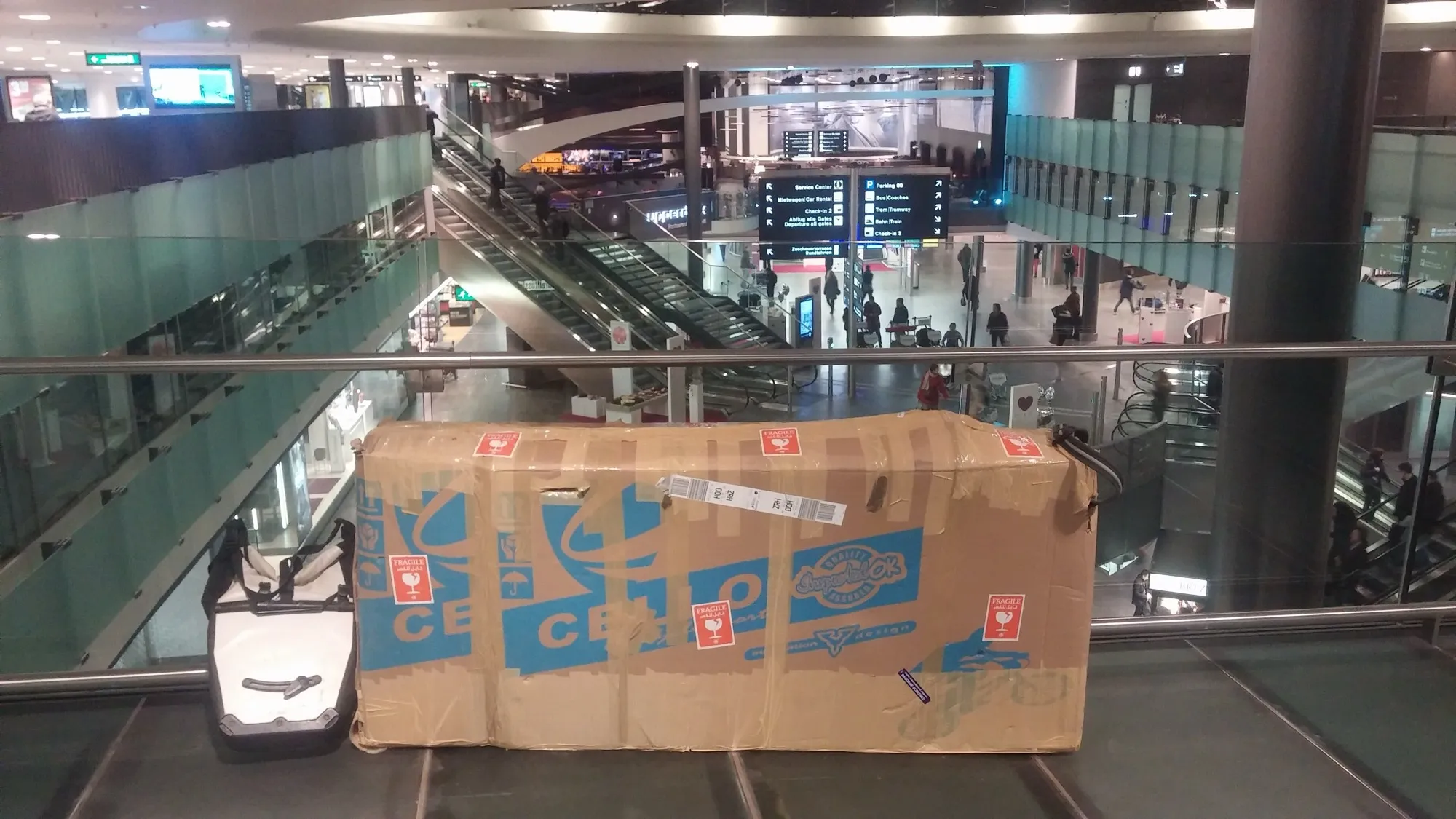 A boxed bicycle in cardboard with fragile stickers, standing beside a pannier in an airport terminal