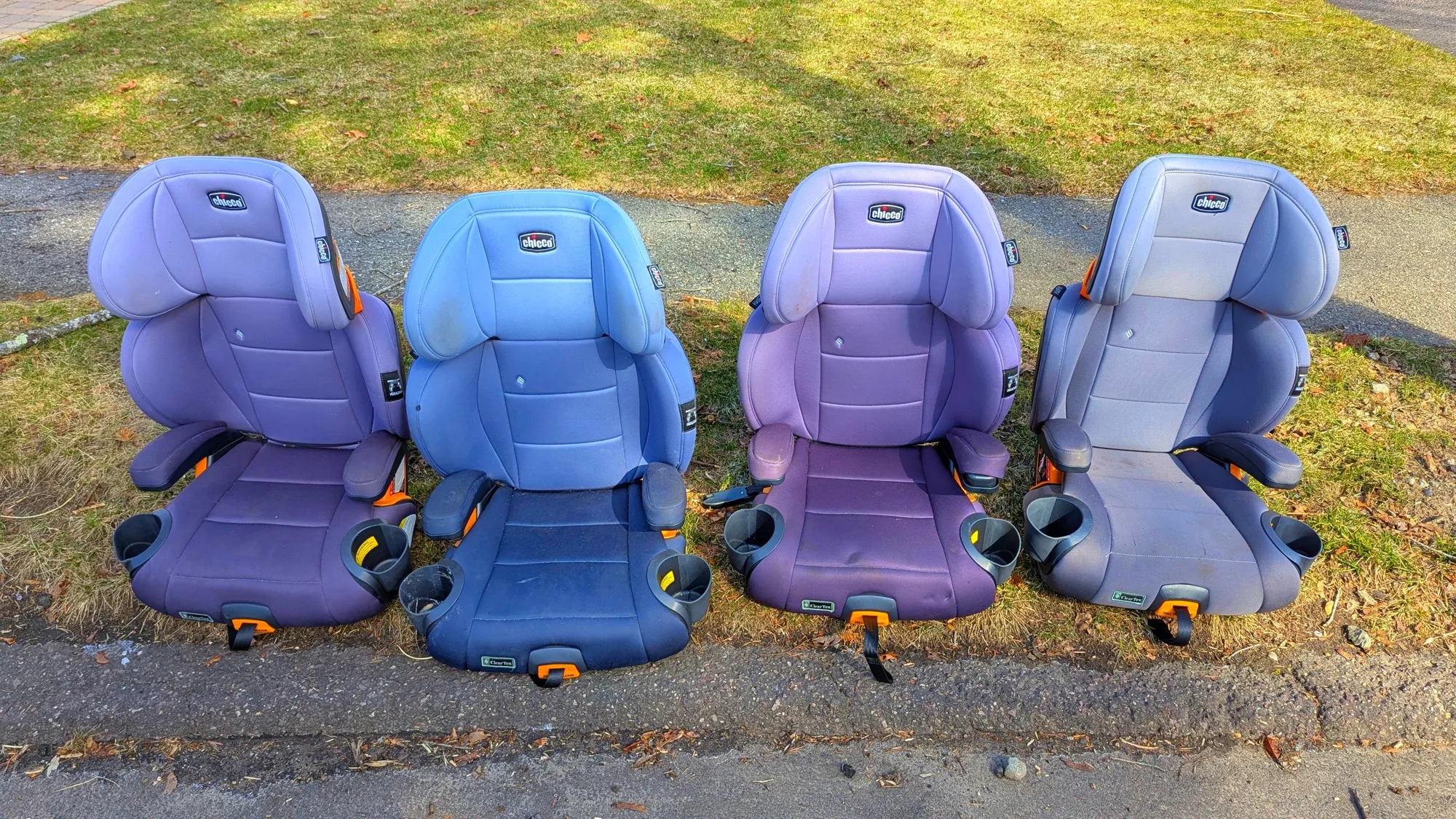 Four high-back child booster seats lined up side by side on a grass verge