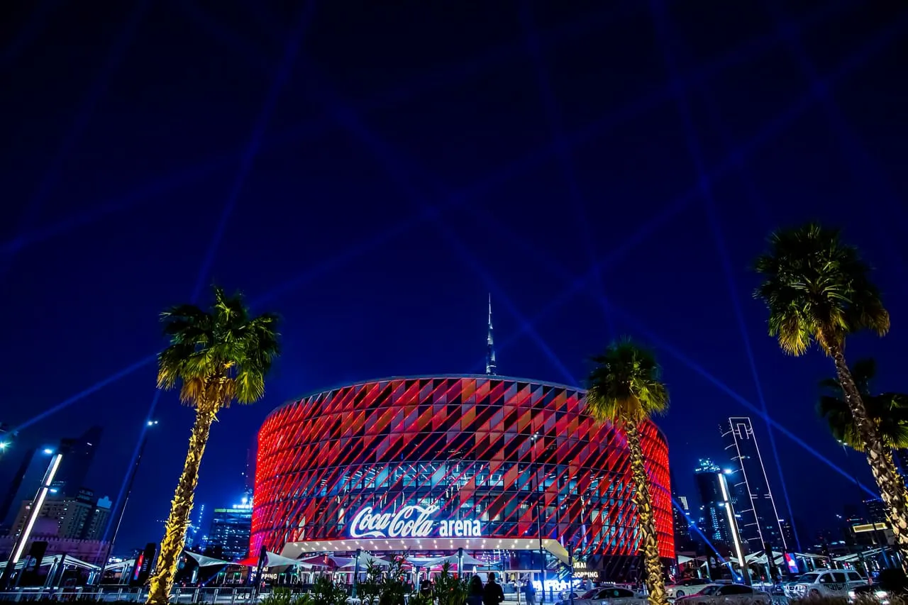 Coca-Cola Arena lit red at night in City Walk, with Burj Khalifa rising behind it