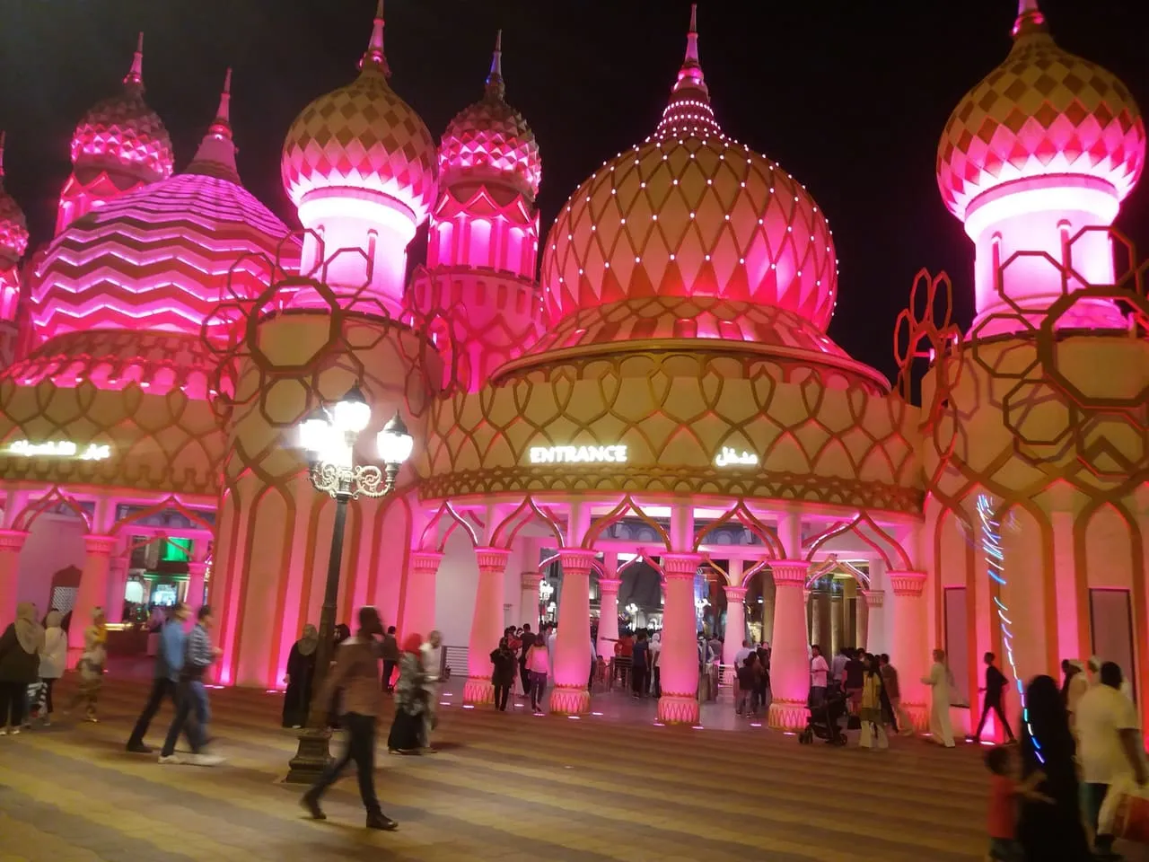 A pink-lit entrance gate at Global Village after dark, with visitors walking in