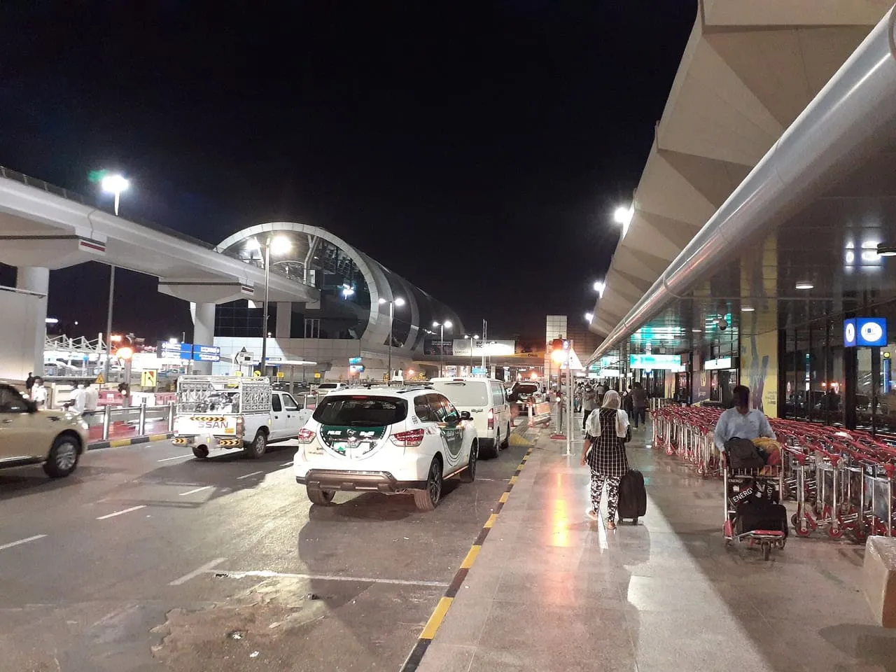 Cars at the kerb outside Terminal 1 of Dubai International Airport at night, with the Airport Terminal 1 metro station behind