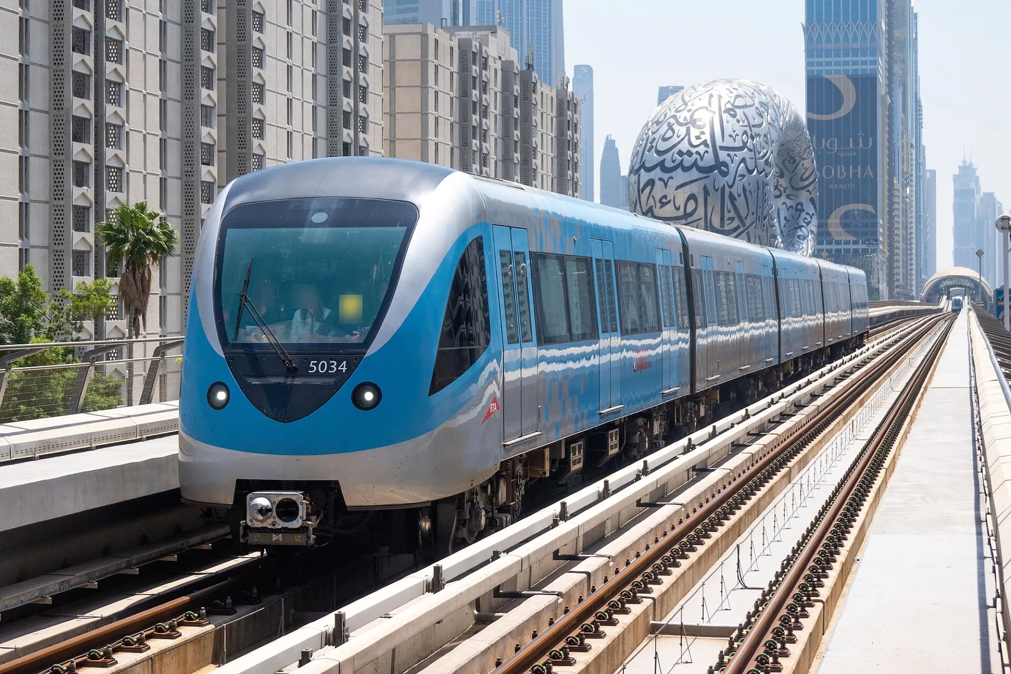 A blue Dubai Metro Red Line train arriving at a raised station with the Museum of the Future behind