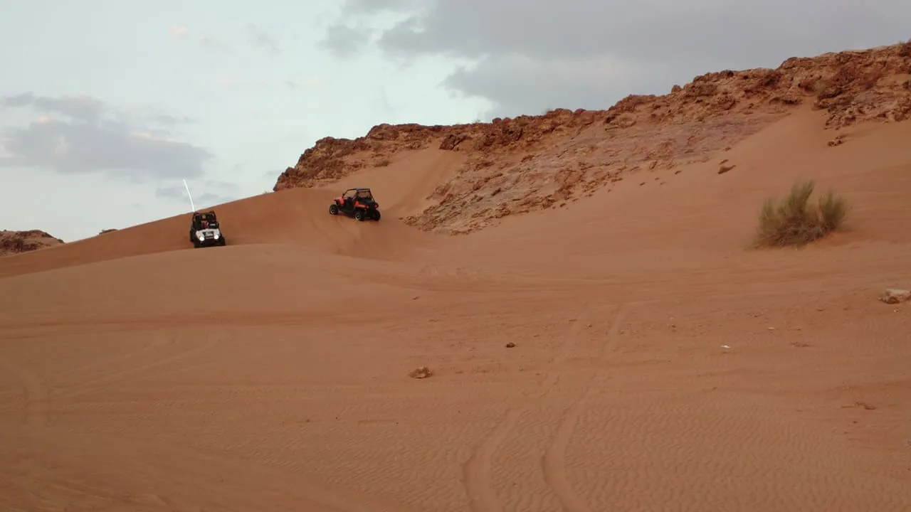 Two dune buggies driving over orange sand below a rocky ridge in the Sharjah desert