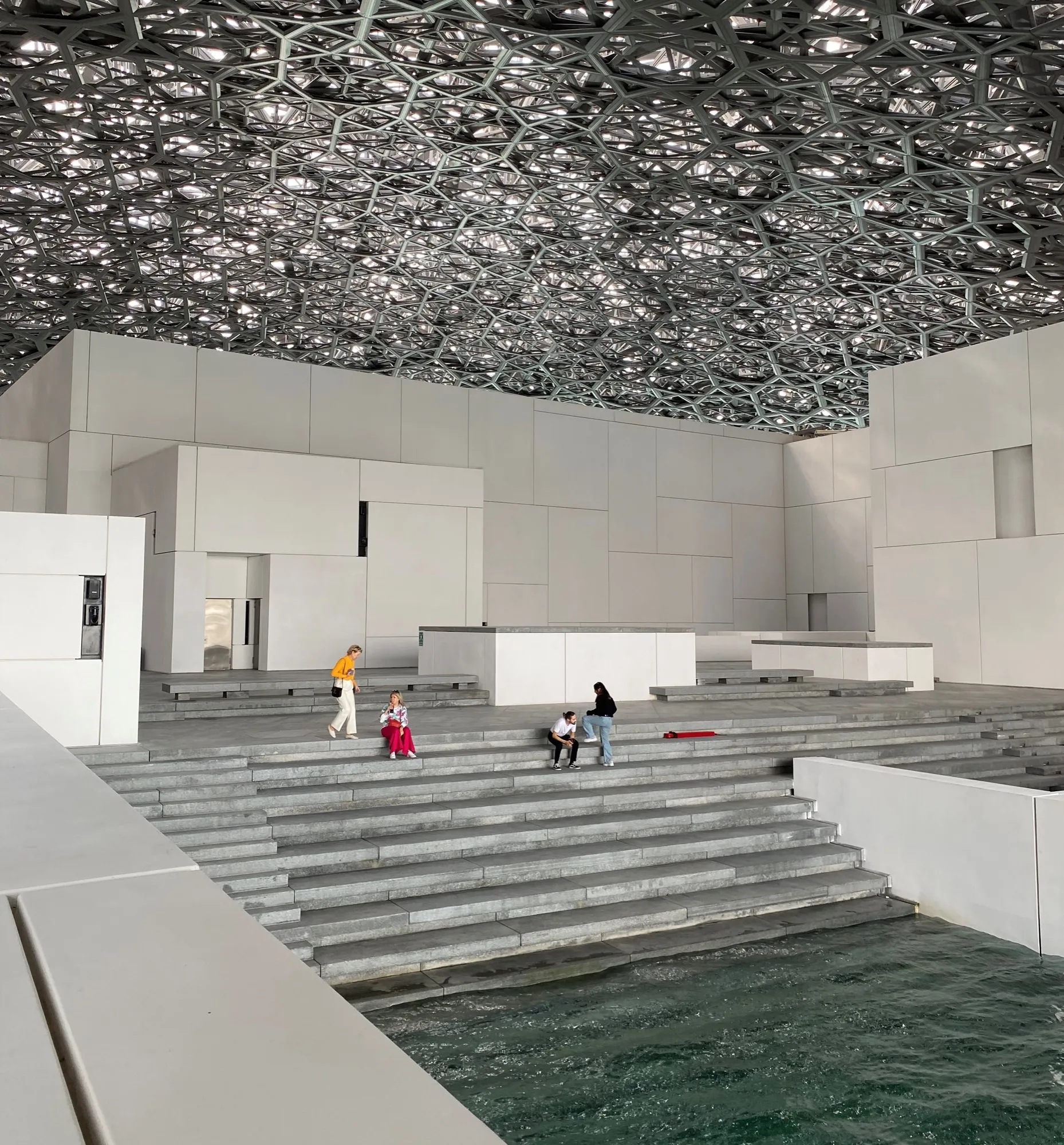 Visitors sitting on stone steps beside the water under the lattice dome of Louvre Abu Dhabi