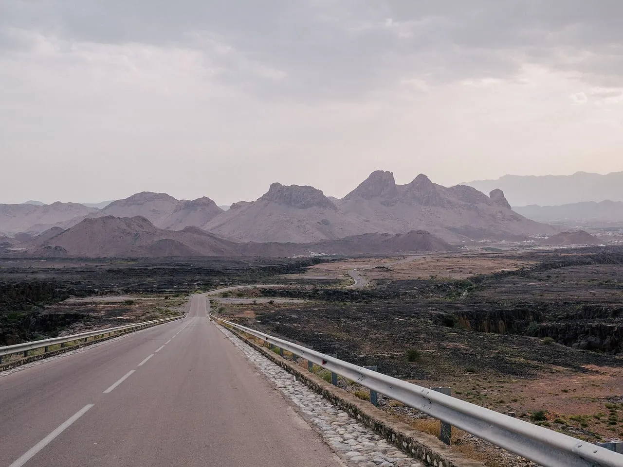 A two-lane road near Al Barida in Ad Dakhiliyah, Oman, running across the plain toward the Hajar Mountains on the way to Jebel Akhdar