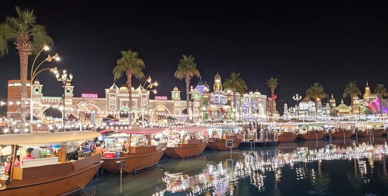 Global Village pavilions lit up at night across the lake, with wooden boats moored in front