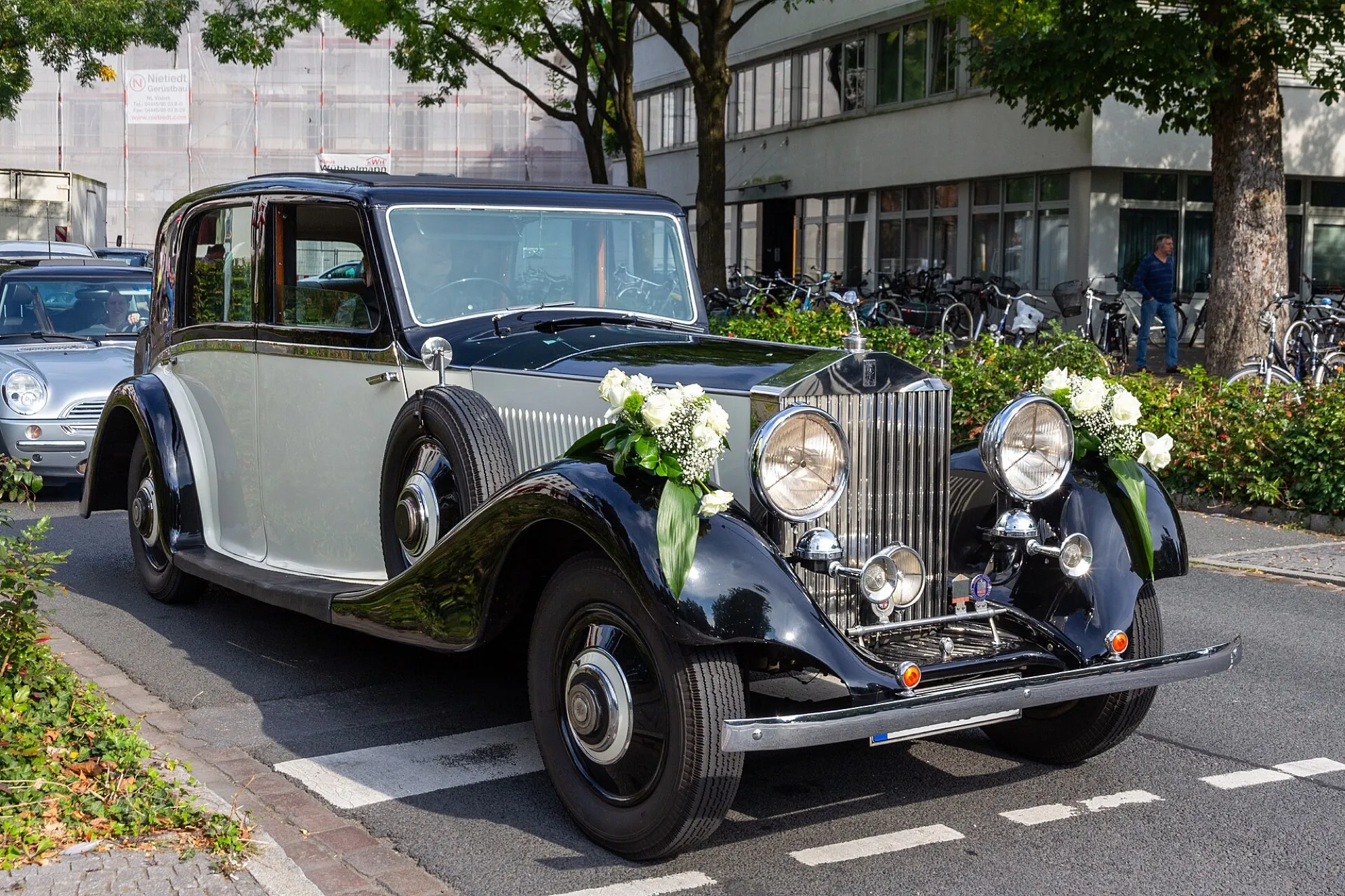 A grey and black vintage Rolls-Royce saloon decorated with white roses on both front wings, used as a wedding car