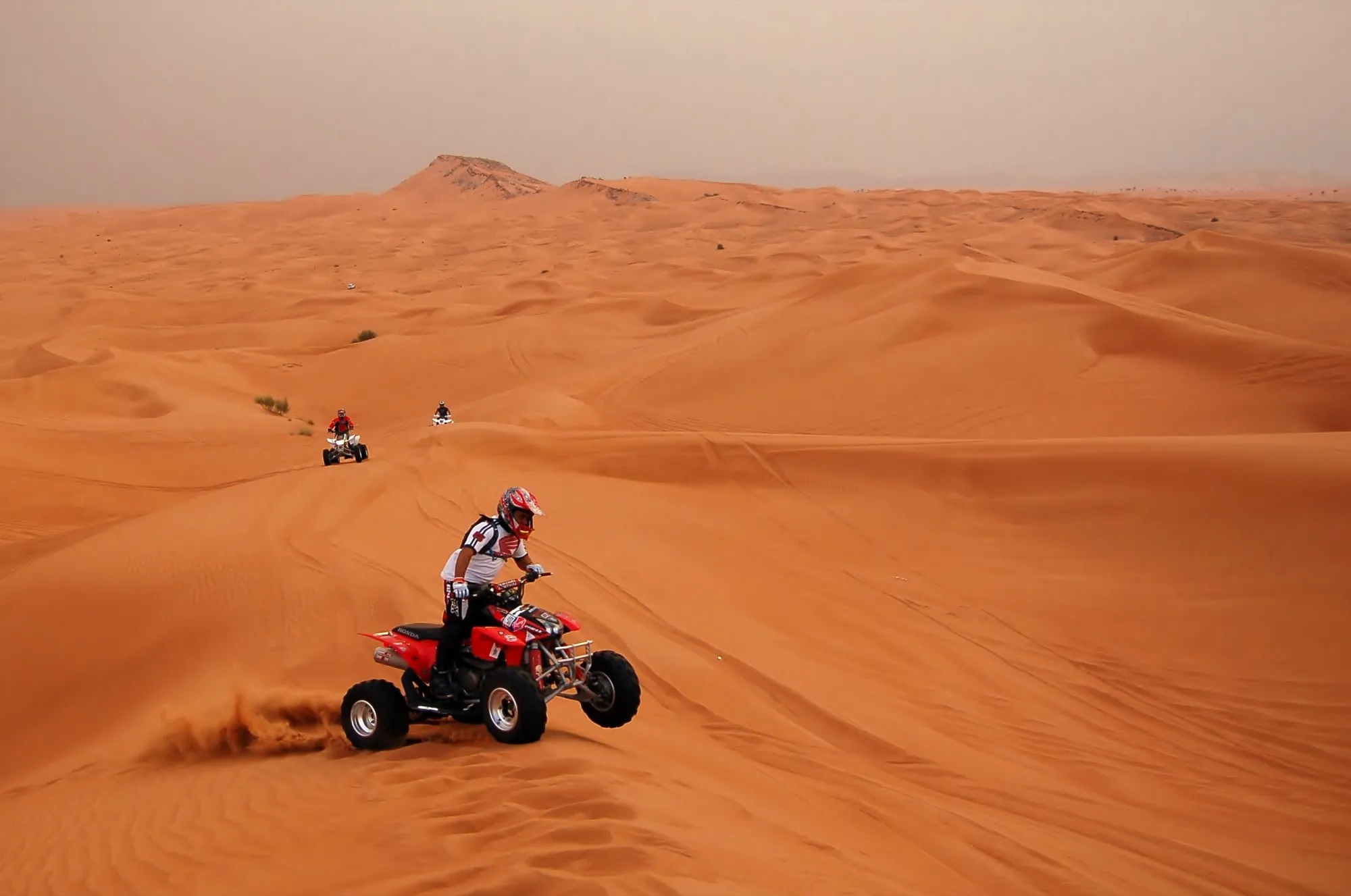 A rider in a helmet on a red quad bike crossing red sand dunes in the Dubai desert, with two more quads behind
