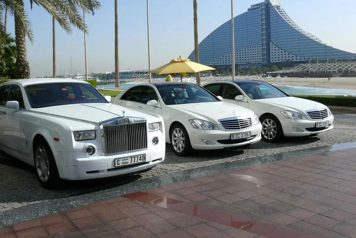 A white Rolls-Royce Phantom and two white Mercedes-Benz S-Class cars with Dubai plates outside the Burj Al Arab, with the Jumeirah Beach Hotel behind