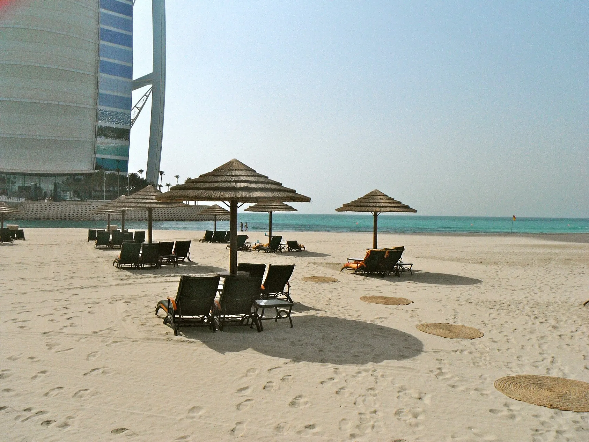 Sun loungers and thatched parasols on a Jumeirah beach in Dubai, with the Burj Al Arab at the left of the frame