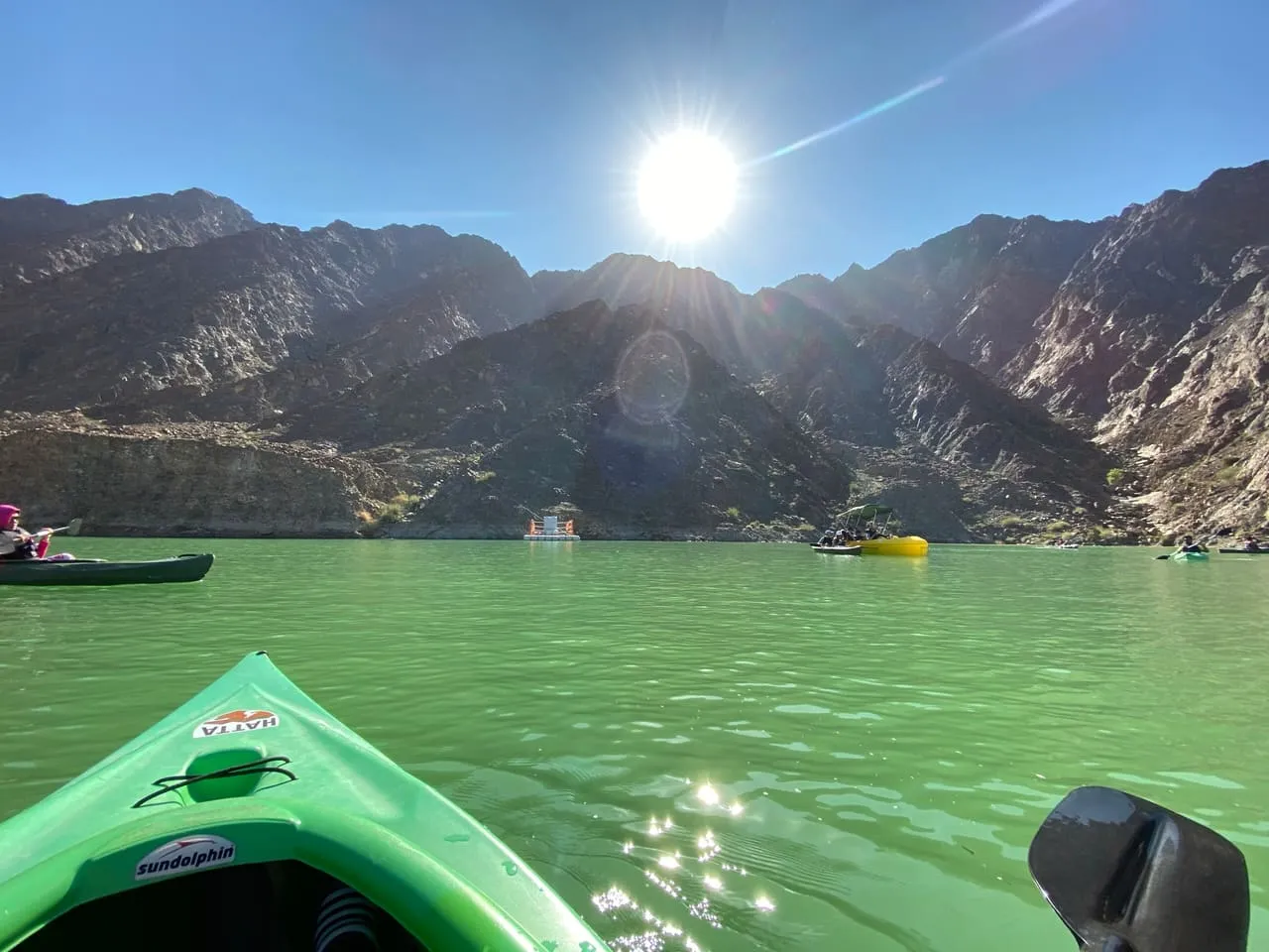 The bow of a green kayak on the water of Hatta Dam, with other kayaks and the Hajar Mountains ahead