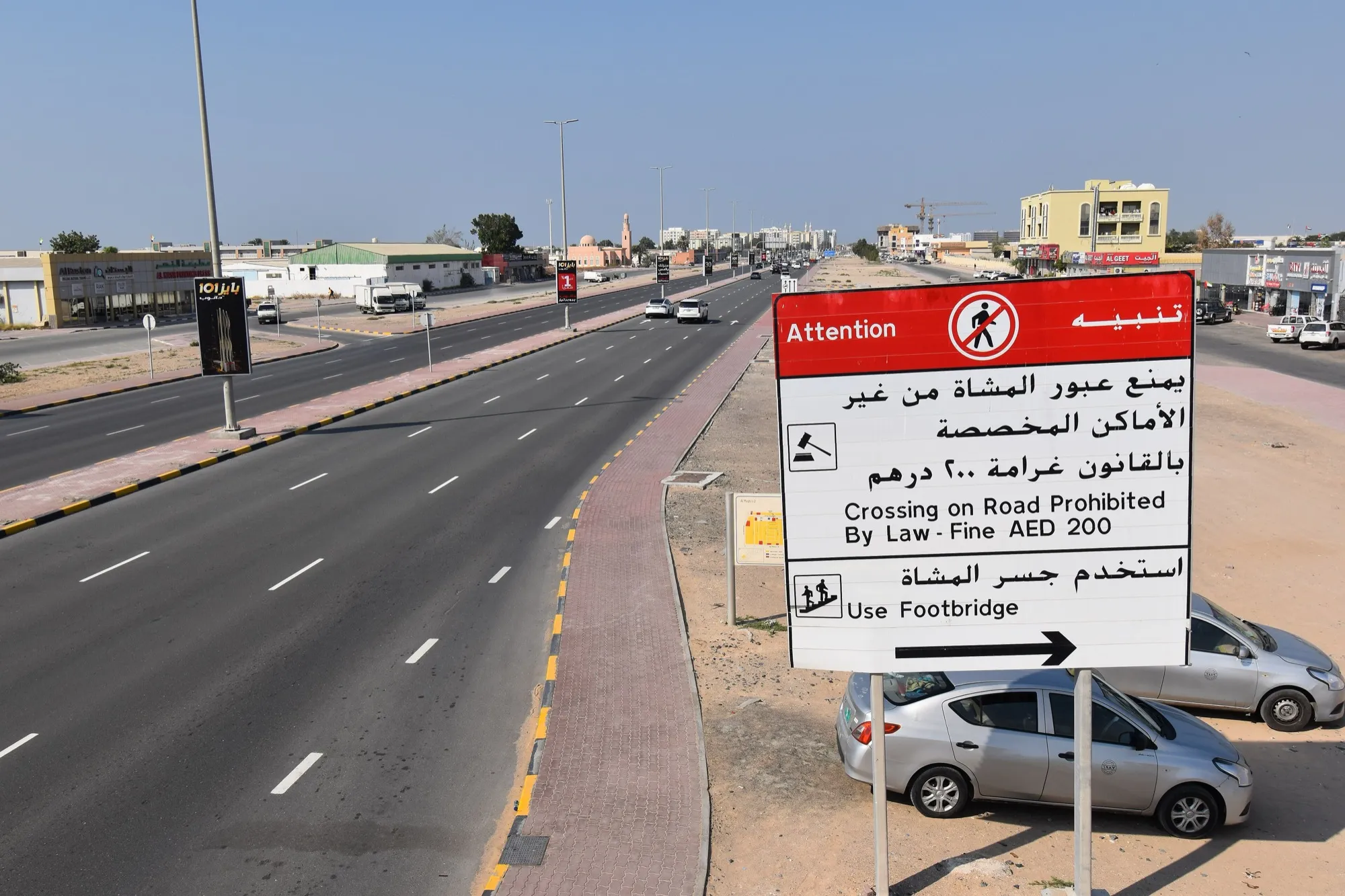 A wide multi-lane road in Umm Al Quwain with shops and a footbridge sign beside it