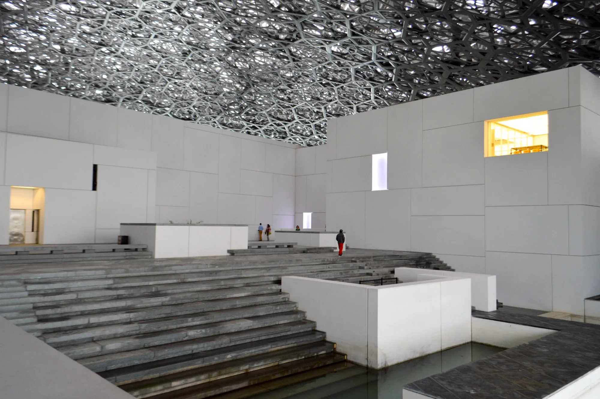 White gallery blocks, stone steps and a pool under the dome at Louvre Abu Dhabi