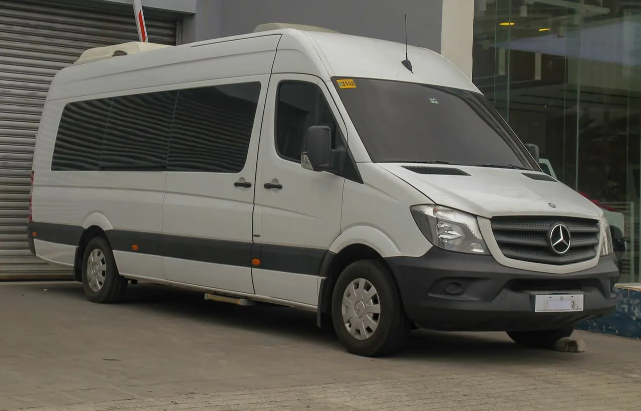 A white high-roof Mercedes-Benz Sprinter minibus with passenger windows along its side