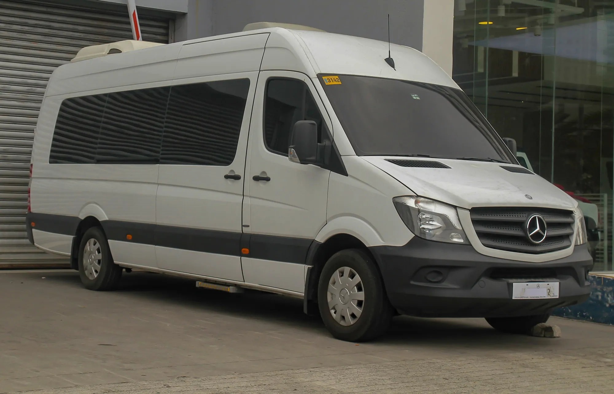 A white high-roof Mercedes-Benz Sprinter minibus with passenger windows along its side