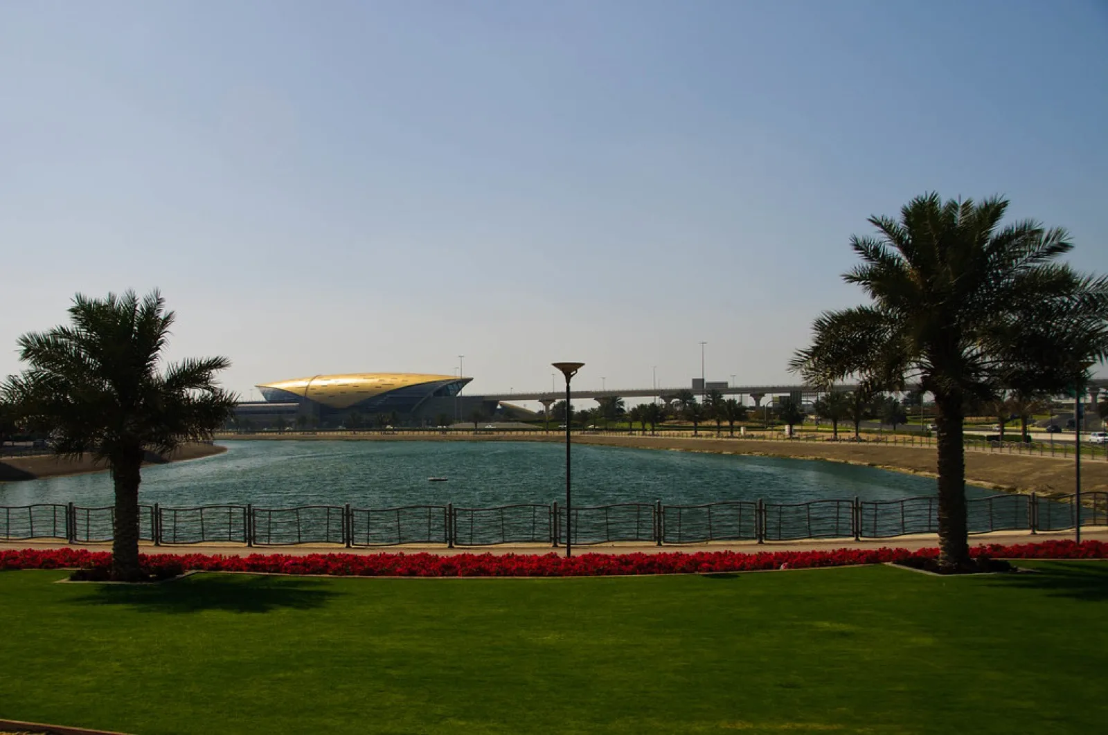 A Dubai Metro station with a golden roof beside a lake, with palms and a lawn in the foreground