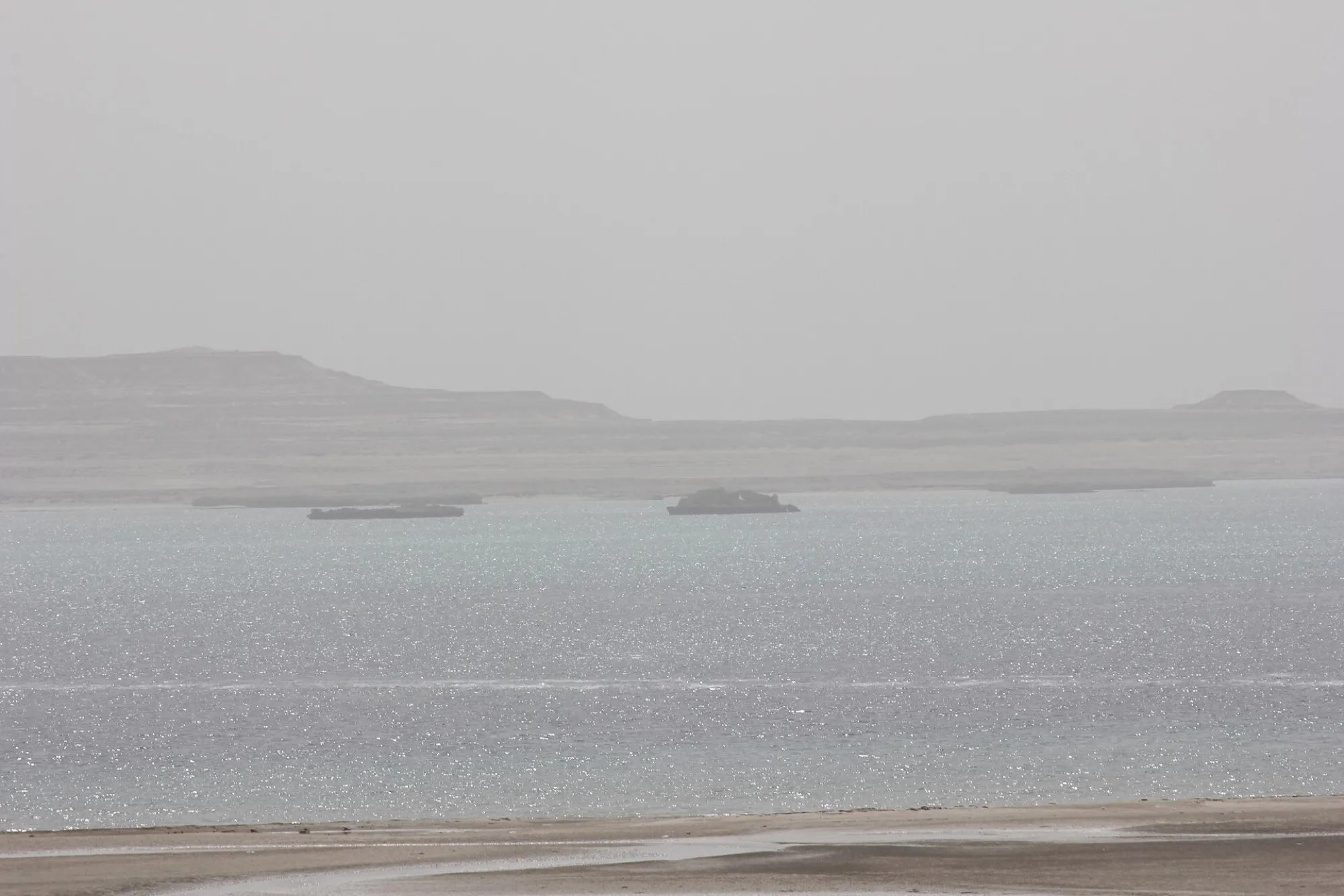 Hazy shore and flat desert hills where Qatar meets Saudi Arabia, with low rock islets in the water