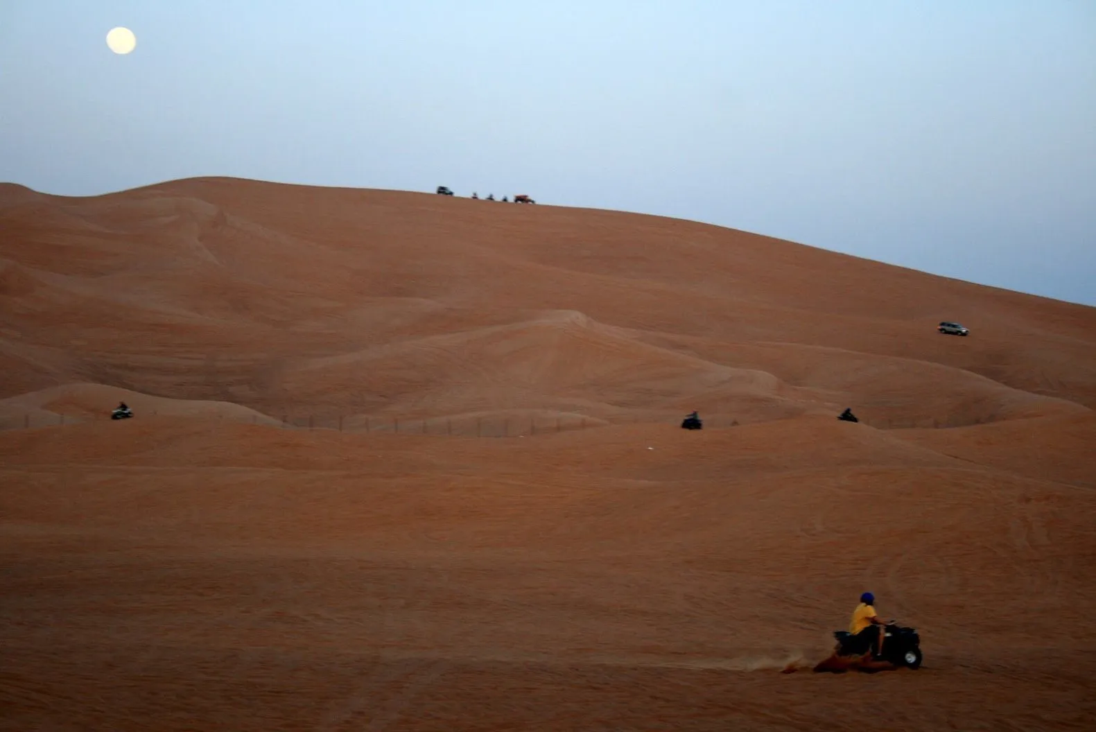 Quad bikes spread across a tall red dune at dusk in the Dubai desert, with a rider in a blue helmet in front