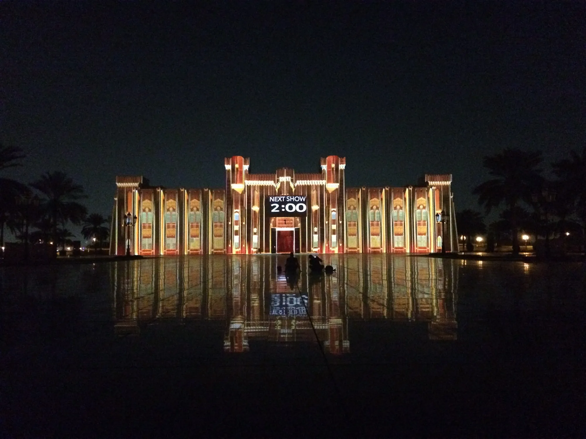 A building facade lit by a projection show at the Sharjah Light Festival, with a next-show board in front
